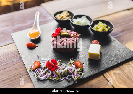 Rindertartar mit rohem Ei und Gemüse auf einer Steinplatte. Hochwinkelschuss. Holztisch als Hintergrund. Hochwertiges Foto Stockfoto