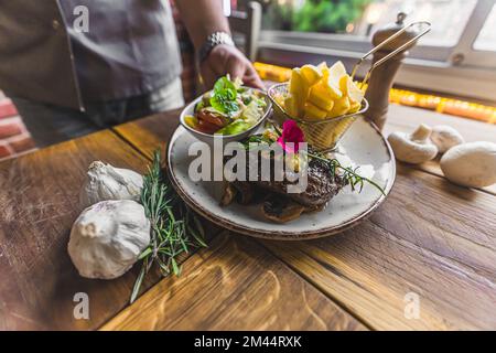 Der Kellner, den man nicht wiedererkennt, hat ein weißes Restaurant mit einem wunderbar servierten Kotelettsteak zusammen mit Salat und Pommes frites serviert. Knoblauch als Dekoration. Holztisch. Hochwertiges Foto Stockfoto