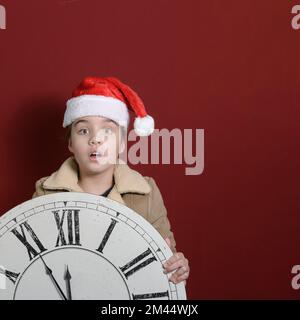 Fünf Minuten bis zum neuen Jahr. Ein Mädchen mit einer großen, alten Wanduhr in der Hand. Quadratisches Format. Er trägt einen Weihnachtsmannmütze, einen begeisterten Blick in die Kamera. Stockfoto