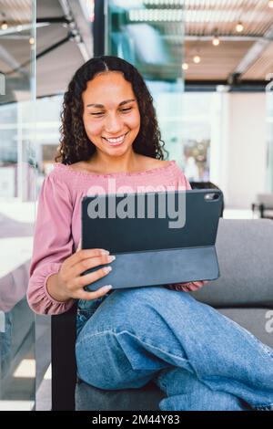 Hispanische Studentin, die lächelt und in ihrer Freizeit vor einem Café auf einem Tablet sitzt Stockfoto