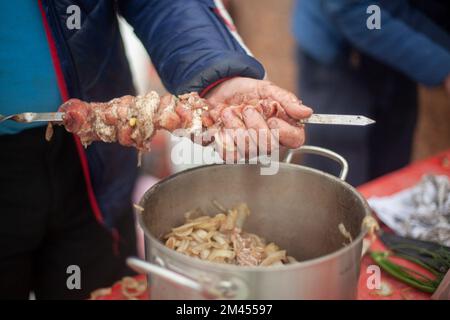 Ich lege Fleisch auf das Messer. Ich koche Fleisch mit den Händen. Straßenküche. Kaukasische folkloristische Küche. Ein Mann mit einem Stück Fleisch und Spieß. Stockfoto