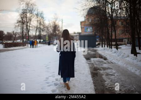 Mädchen geht im Winter die Straße entlang. Frau im Winter in der Stadt. Schlechte Straße im Hof in Russland. Der Student kehrt nach Hause zurück. Stockfoto