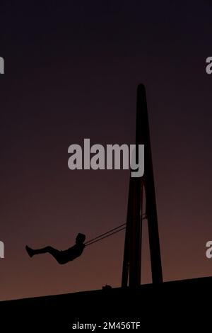 Ein Mann sitzt auf einer Schaukel gegen das Licht in einer Sonnenuntergangssilhouette des Mannes Stockfoto