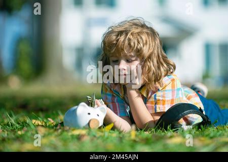 Der Junge spart Geld für den Kauf, hält das rosa Sparschwein, legt sich auf Gras. Kindererziehung zur Berechnung des persönlichen Budgets, Finanzverwaltung und Investitionsförderung Stockfoto