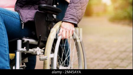 Die Räder drehen sich weiter. Eine nicht wiedererkennbare ältere Frau im Rollstuhl draußen in einem Park während des Tages. Stockfoto