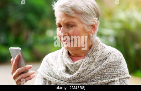Ich habe eine Nachricht. Eine fröhliche ältere Frau, die auf ihrem Handy SMS schreibt, während sie draußen auf einer Bank in einem Park sitzt. Stockfoto