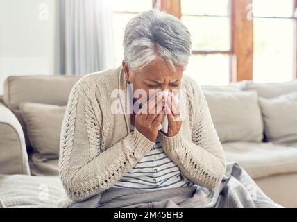 Ich muss jetzt zum Arzt. Eine kranke ältere Frau, die sich mit einem Taschentuch die Nase putzt, während sie auf dem Sofa zu Hause sitzt. Stockfoto