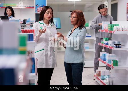 Mediziner erklärt dem Kunden verschreibungspapier, spricht über Medikamente, die auf Apothekenregalen verteilt sind. Apotheker, der Kunden mit Arzneimitteln behilflich ist. Stockfoto