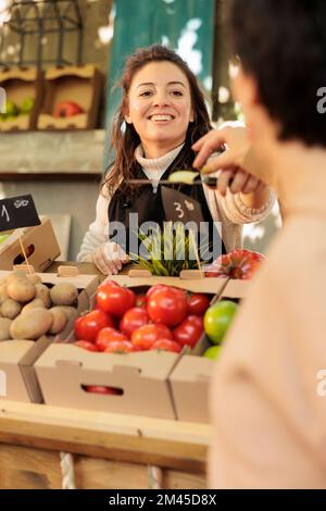 Lächelnder, freundlicher Besitzer eines Bio-Obst- und Gemüsehandels, der dem Kunden auf dem Lebensmittelmarkt verschiedene Äpfelsorten anbietet. Positive Landwirtinnen, die frische einheimische Erzeugnisse verkaufen Stockfoto