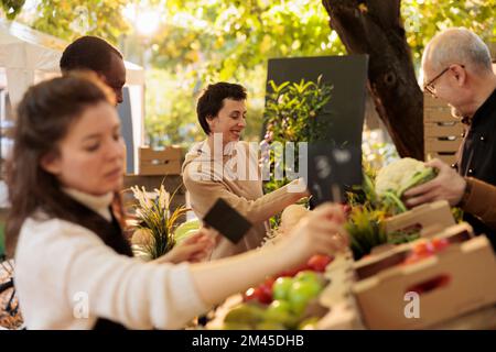 Junge, fröhliche und fröhliche Frau, die das Einkaufserlebnis auf dem lokalen Bauernmarkt genießt, weibliche Kundin, die an einem Stand für frische Bio-Produkte steht und gesunde Produkte aus der Region kauft Stockfoto