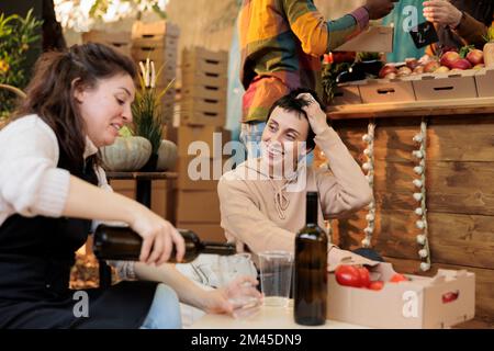 Zwei glückliche Personen beim Food Festival, die hausgemachten Wein probieren, sitzen neben dem lokalen Bauernmarkt draußen. Kunde und Verkäufer, der eine natürliche Flasche Bio-Wein und frische Bio-Produkte trinkt. Stockfoto