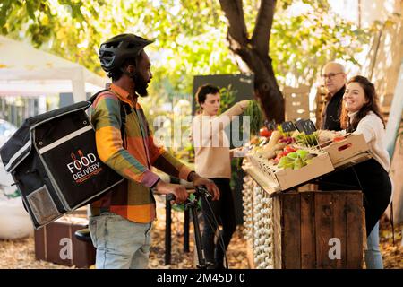 Ein junger afrikanischer Kurier, der einen Lieferrucksack trägt und auf Bestellung auf dem lokalen Lebensmittelmarkt wartet. Landwirte liefern frische Bio-Früchte und -Gemüse an Kunden, um sich mit der neuen Realität vertraut zu machen Stockfoto