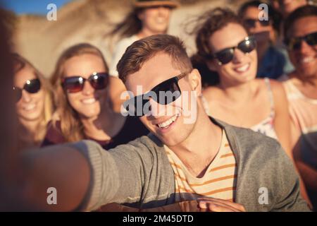 Machen Sie ein Selfie und lassen Sie die guten Zeiten Rollen. Abgeschnittenes Porträt eines gutaussehenden jungen Mannes, der mit seinen Freunden am Strand ein Selfie macht. Stockfoto