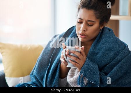 Ich muss mich warm halten. Eine attraktive junge Frau, die sich mit einer Decke bedeckte, während sie zu Hause Kaffee trank. Stockfoto