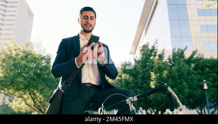 Er schickt ein paar Nachrichten auf dem Weg zur Arbeit. Ein kleiner Winkel von einem gutaussehenden jungen Geschäftsmann, der sein Handy benutzt, während er mit seinem Fahrrad zur Arbeit pendelt. Stockfoto