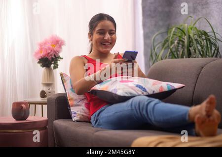 Glückliches Teenager-Mädchen, das Handy benutzte, während es sich auf dem Sofa im Wohnzimmer entspannte Stockfoto