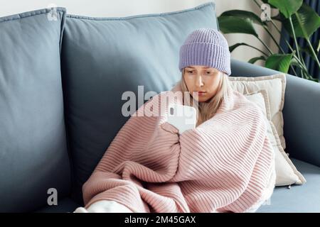 Unglückliche junge Frau Influencer in Decke eingewickelt mit Wollmütze sitzen auf der Couch, schauen Smartphone und fühlen sich kalt. Energiekrise in Europa fällig Stockfoto