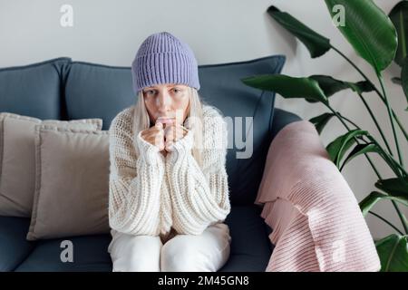 Unglückliche junge Frau, die zu Hause warme Kleidung trägt und sich kalt fühlt. Energiekrise in Europa infolge von Inflation und Krieg. Erhöhung des Preises für Naturprodukte Stockfoto