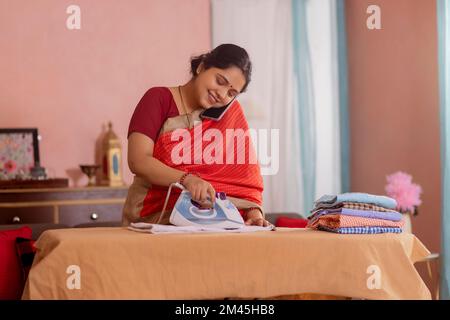 Porträt einer glücklichen Frau, die mit dem Handy telefoniert, während sie zu Hause Kleidung bügelt Stockfoto