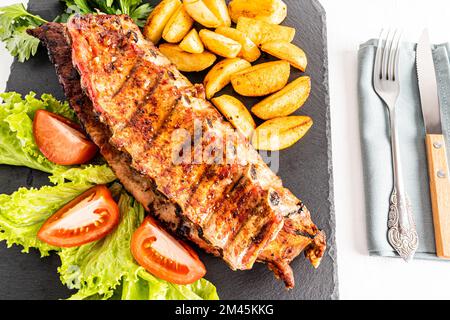 Blick von oben auf die köstlichen saftigen gegrillten Schweinerippchen mit Kartoffelscheiben und Salat auf einem Schieferbrett. Besteck. Köstliches Essen Stockfoto