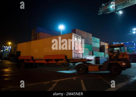 Hafen-Container-Terminal bei Nacht. Bewegungsunschärfe eines Containers auf einem Traktor im Vordergrund. Stockfoto