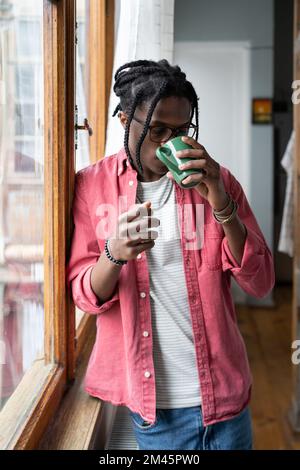 Aufgebrachter Afrikaner, der neben dem Fenster steht und eine Tasse Tee trinkt, um sich zu beruhigen und Stress zu reduzieren Stockfoto