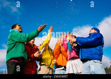 Multiethnische Gruppe junger, glücklicher Freunde, die sich im Winter im Freien treffen und eine Party mit Konfetti-Shooter feiern, Winterjacken tragen und Spaß haben Stockfoto