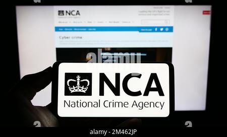 Person, die ein Mobiltelefon mit dem Logo der British National Crime Agency (NCA) auf dem Bildschirm vor der Webseite hält. Konzentrieren Sie sich auf das Display des Telefons. Stockfoto