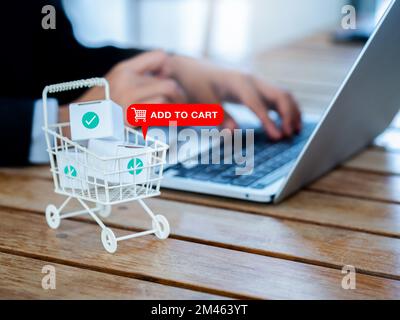 Das Symbol zum Warenkorb hinzufügen wird auf dem weißen Einkaufswagen angezeigt. Der Warenkorb enthielt Paketkästchen mit Häkchen in der Nähe des Geschäftsmanns, der einen Laptop verwendet Stockfoto