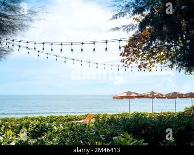 Seascape. Sommerblick mit festlichen hängenden Glühbirnen, orange gestreiften Sonnenschirmen, Sonnenliegen unter den Bäumen am Strand, Sand am blauen Himmel Stockfoto