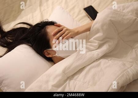 Kranke Frau, die eine Maske im Bett trägt, ihre Hände über den Augen, Handy auf dem Bett, Mädchen, das im Bett schläft Stockfoto