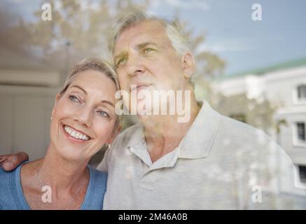 Umarmen, denken und Seniorenpaar an einem Fenster mit Zukunftsvision, Ruhestandsruhe und Entspannen in ihrem Zuhause. Idee, Liebe und ältere Männer und Frauen Stockfoto