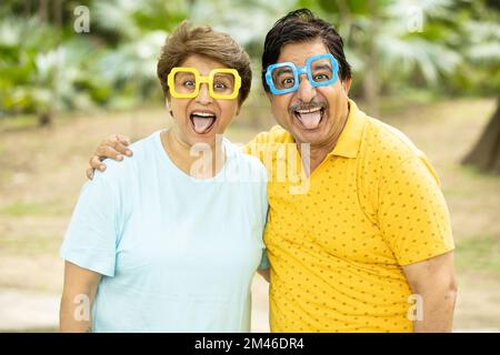 Portrait eines verrückten, aufgeregten, lustigen indischen Seniorenpaares, das im Park eine flippige Brille trägt. Reife Menschen machen Gesichter. Stockfoto