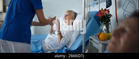 Junge Krankenschwester gibt dem Seniorpatienten auf der Krankenhausstation ein Glas Wasser. Stockfoto