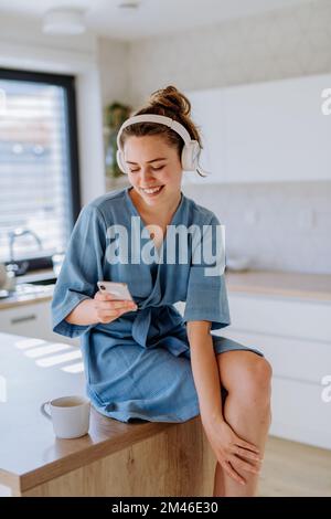 Junge Frau, die Musik hört, mit dem Telefon scrollt und morgens in ihrer Küche Kaffee trinkt. Stockfoto