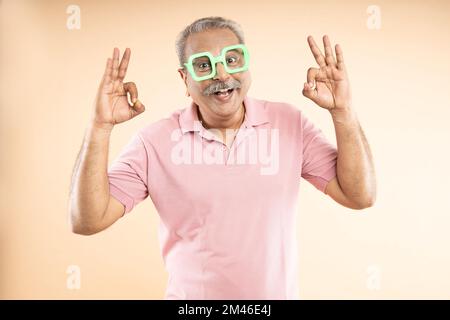 Ein glücklicher Senior-indianer, der eine funky Sonnenbrille trägt und isoliert auf beigefarbenem Hintergrund steht. Pensionierungsleben. Stockfoto