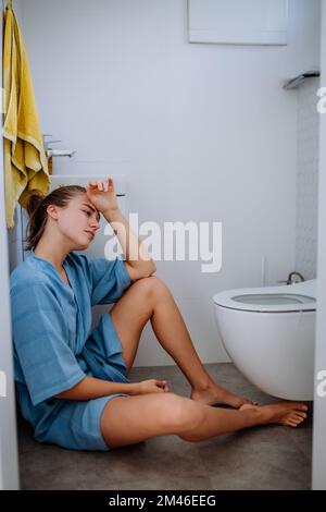 Junge kranke Frau, die auf dem Boden neben der Toilette sitzt. Stockfoto