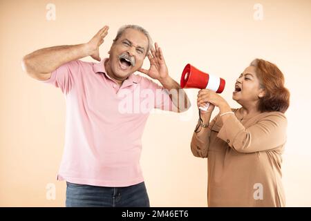 Ältere indianerin schreit oder schreit einen Mann in Megaphon isoliert auf beigem Hintergrund an. Verkünden Sie die Rabatte. Asiatische Ehefrau ärgert Ehemann. Stockfoto