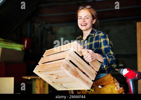 Frau mit Holzkiste in der Hand und lächelnde Frau in einer Tischlerwerkstatt. Stockfoto