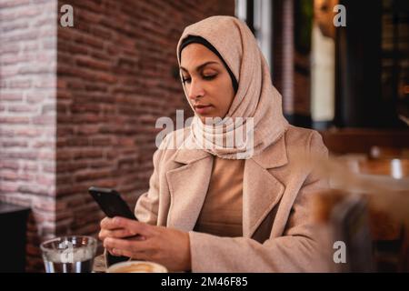 Junge muslimische Frau, die im Café sitzt und mit dem Smartphone scrollt. Stockfoto