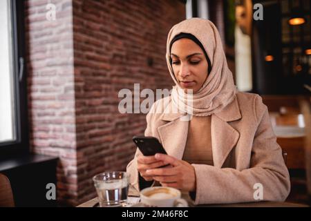 Junge muslimische Frau, die im Café sitzt und mit dem Smartphone scrollt. Stockfoto