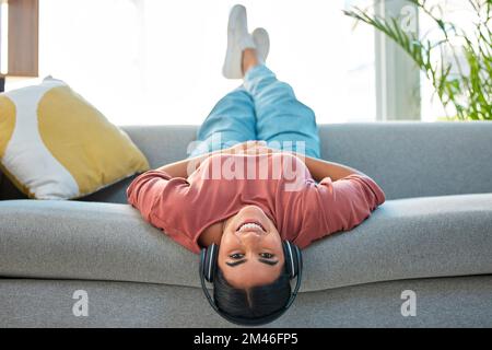 Frau, Sofa und entspannen Sie sich mit Kopfhörern, Musik und einem Lächeln im Porträt, während Sie kopfüber in der Lounge sitzen. Glückliche Dame, Couch und Lügen für Glück, Streaming Stockfoto