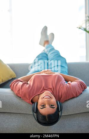 Frau, Sofa und kopfüber mit Kopfhörern, Lächeln oder Musik im Porträt zum Entspannen in der Lounge. Glückliche Dame, Sofa oder Audio für Glück, Streaming Stockfoto