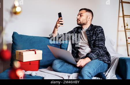 Ein erwachsener Mann verwendet sein Mobiltelefon und seinen Laptop, während er zu Hause in den Weihnachtsferien auf der Couch sitzt. Stockfoto