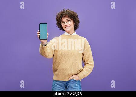 Junge, brünette Frau, die ihr Smartphone mit einem leeren blauen Bildschirm hält, während sie vor lila Hintergrund steht Stockfoto