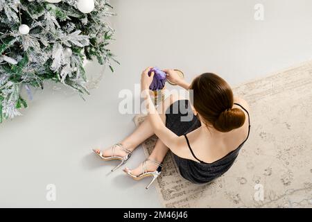 Blick von oben auf eine Frau in schwarzem glitzerndem Kleid, die auf dem Boden neben einem Weihnachtsbaum sitzt und einen violetten Seidenschal aus einer Geschenkbox nimmt Stockfoto