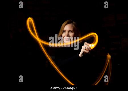 Schreiben Sie mit Licht bei langer Belichtung im Dunkeln. Eine Frau mittleren Alters malt mit einer Taschenlampe. Stockfoto