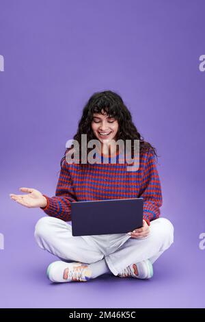 Glückliches Mädchen mit dunklem, langen, gewelltem Haar, das in einem Video-Chat kommuniziert, während es isoliert vor der Kamera sitzt und ihr Notebook auf den Knien liegt Stockfoto