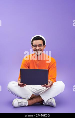 Erfolgreicher junger männlicher Designer oder Programmierer, der ein Notebook verwendet und auf die Kamera schaut, während er mit überkreuzten Beinen auf dem Boden sitzt Stockfoto