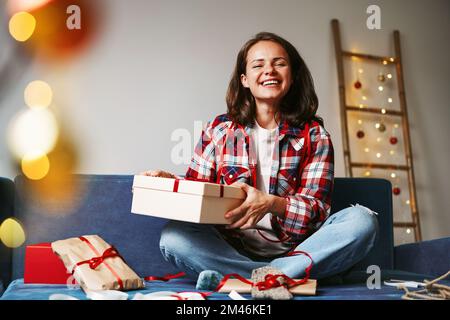 Junge, brünette Frau im karierten Hemd, die auf dem Sofa sitzt und ein Weihnachtsgeschenk in der Hand hält und lächelt. Stockfoto